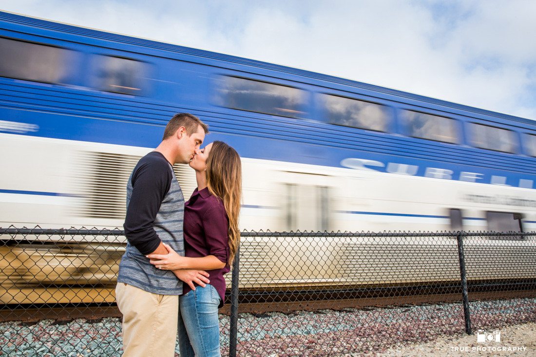 Young couple kissing in front of a moving train for their engagement photo shoot in Del Mar, California.