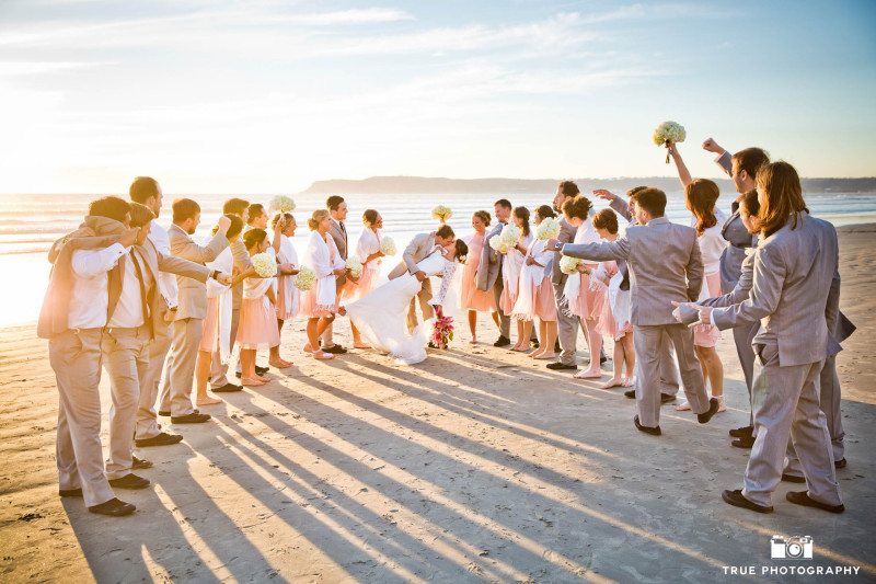 Bridal party cheers while wedding couple dip and kiss on Coronado beach