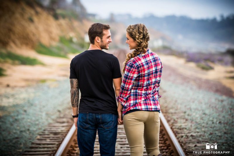 Couple holding hands looking at each other on train tracks.