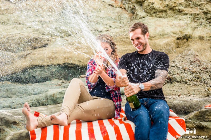 Fun couple popping champagne during engagement session at the beach.