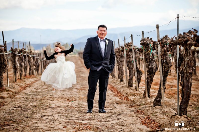 Groom standing in vineyard poses while daughter jumps in background