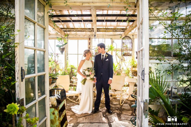 Bride and Groom hold hands and romantically look at one another in rustic greenhouse shed at ranch wedding