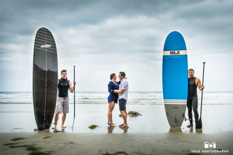 Engaged couple kiss on beach between two surfers