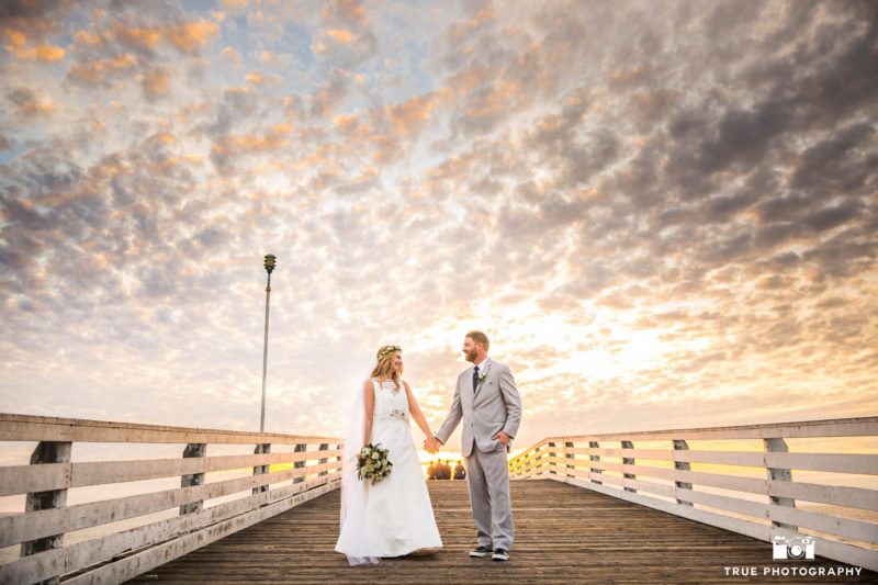 Bride and groom walk down pier at sunset.