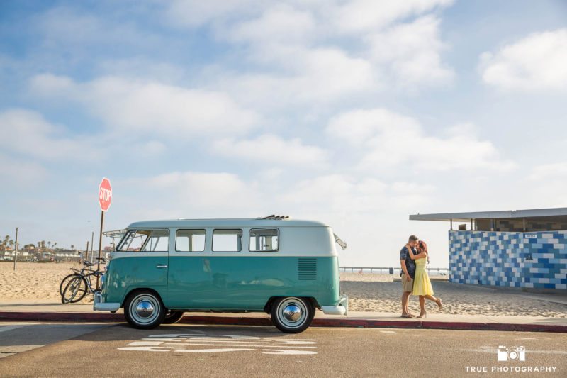 Engaged couple embrace and kiss next to vintage Volkswagen Bus at beach