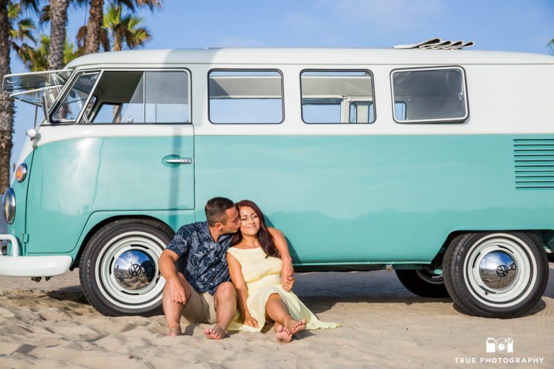 Engaged couple sit against vintage Volkswagen bus on sandy beach during engagement session