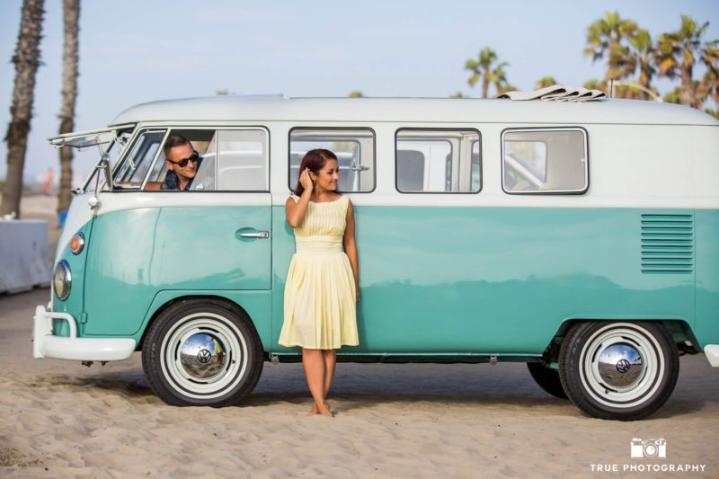 Bride-to-be stands outside vintage Volkswagen bus while Groom-to-be looks at her and drives on beach