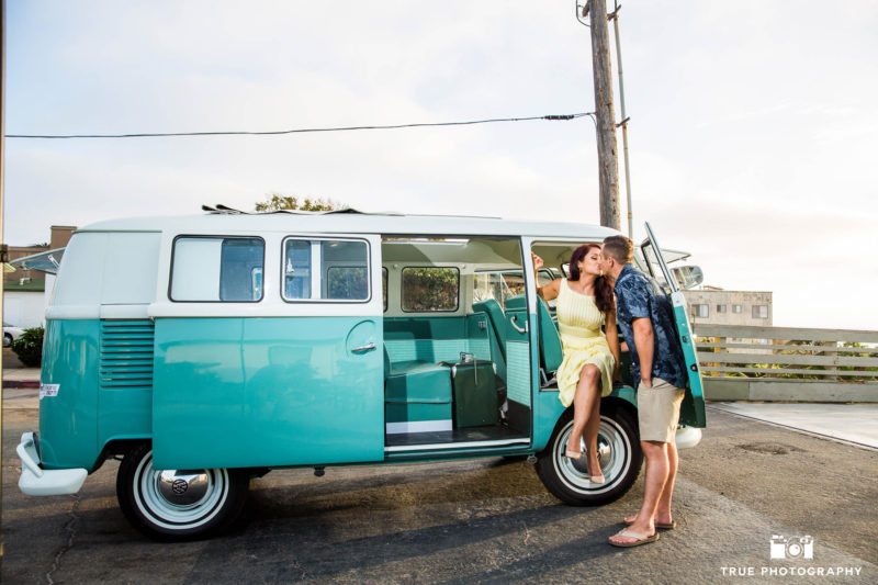 Engaged couple kiss in passenger door of vintage volkswagen bus at beach