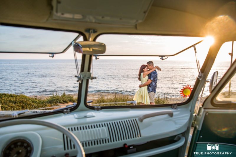 Unique photo of engaged couple embracing during sunset through window of vintage Volkswagen bus during beach engagement session