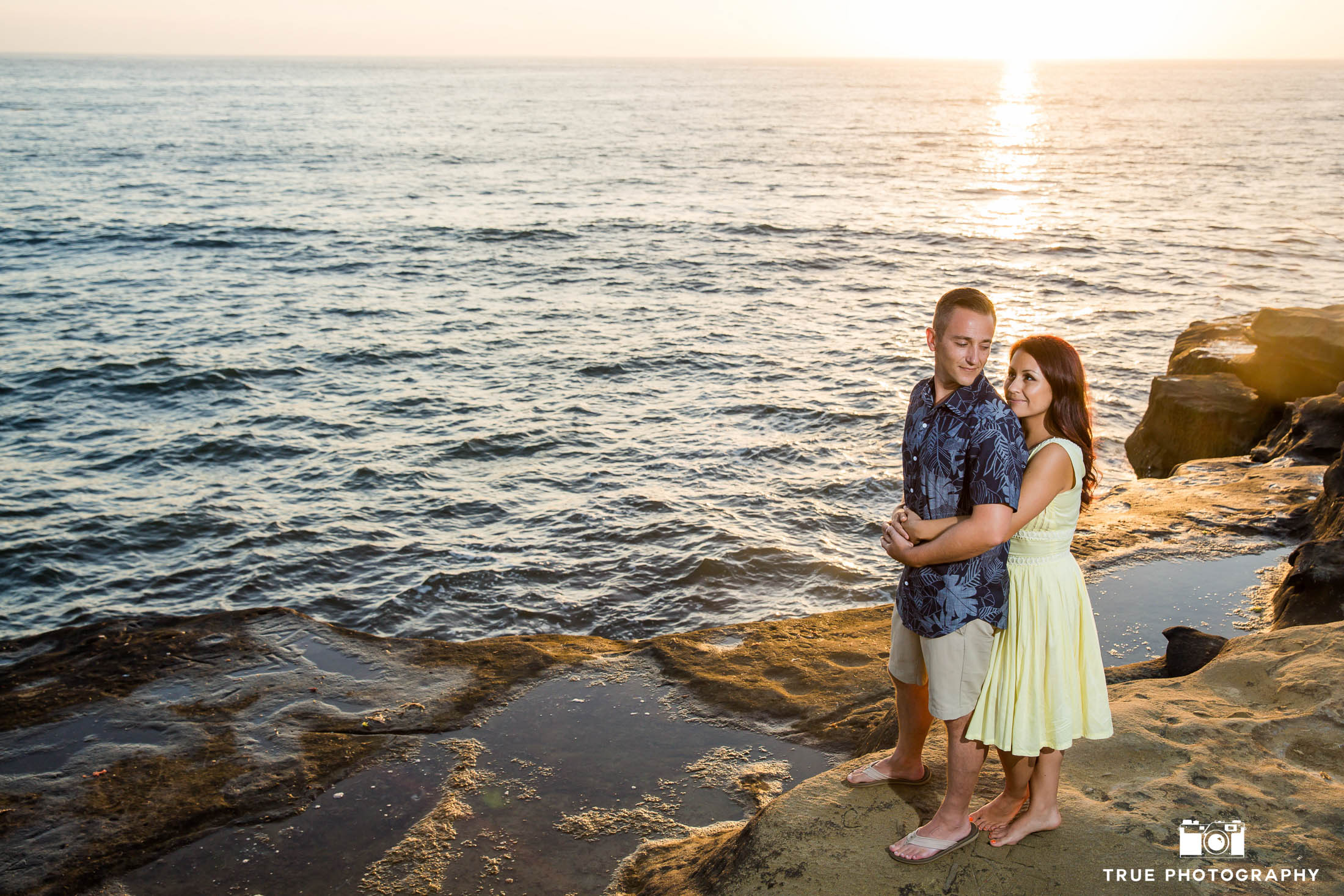 Engaged couple embrace and look at one another on rocks overlooking ocean during beach engagement session