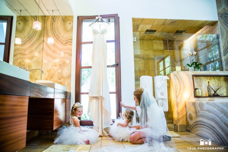 Bride sitting on floor of bathroom with flowergirls and wedding dress