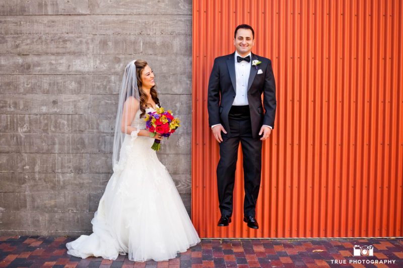 Funny portrait of groom jumping while bride looks on and laughs
